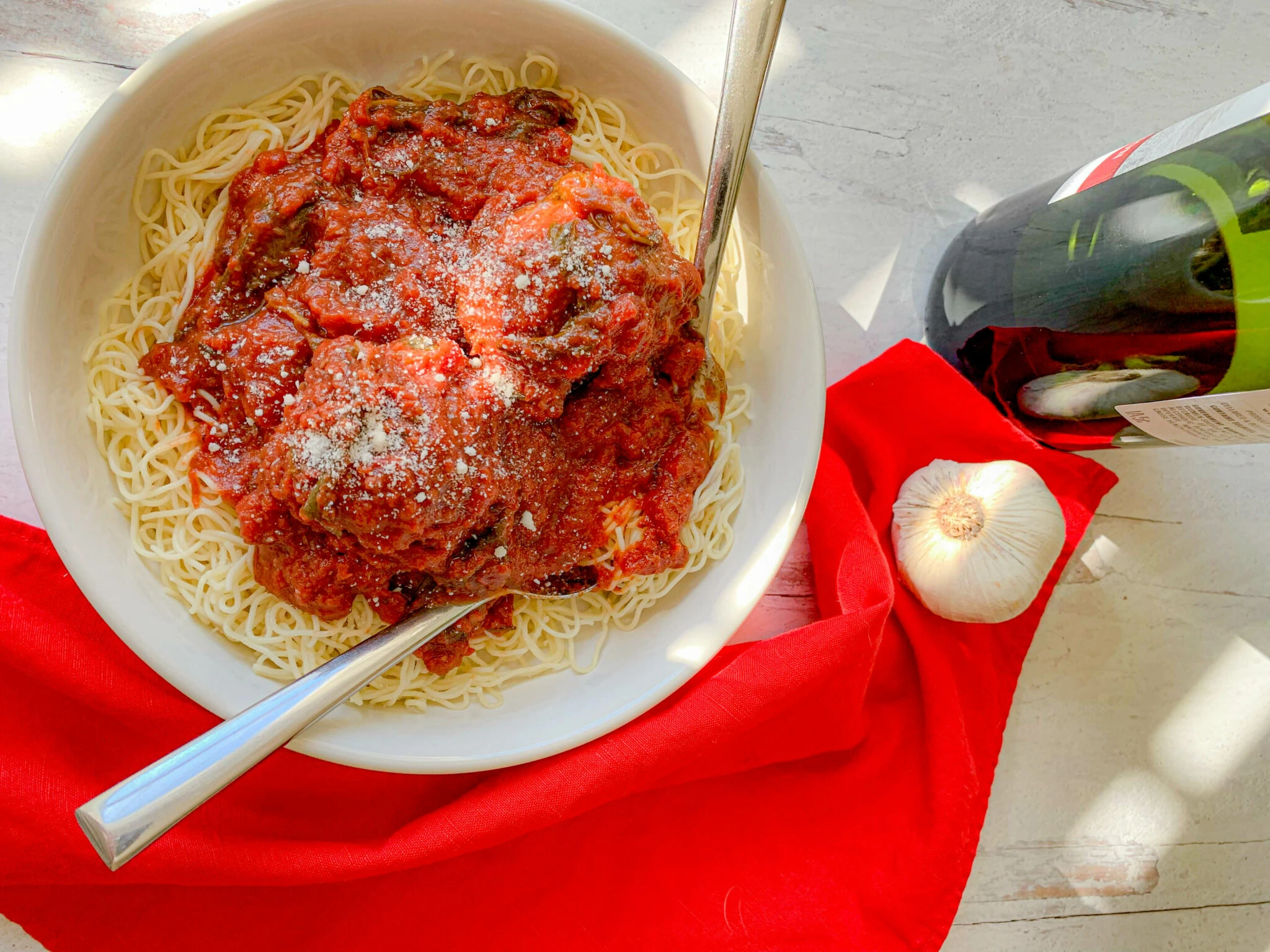 A bowl of spaghetti topped with marinara sauce and grated cheese is on a white surface. A fork is in the bowl. Next to the bowl, there is a red cloth, a whole garlic bulb, and a bottle of wine. The scene is well-lit, enhancing the vibrant colors of the food and props.