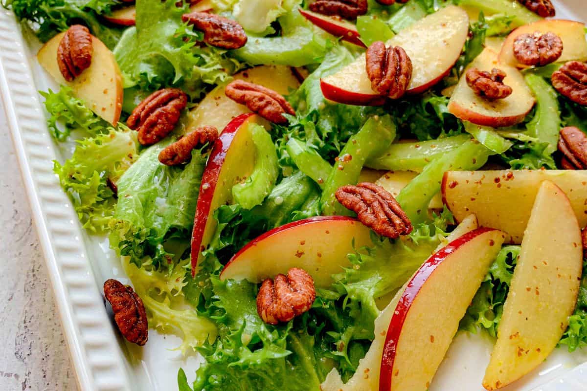 A fresh salad with sliced apples, pecans, and mixed greens on a white rectangular plate. The apples have a red peel and are thinly sliced, while the greens provide a leafy background, creating a colorful and healthy dish.