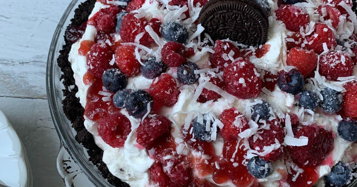 A dessert topped with raspberries, blueberries, shredded coconut, and a chocolate cookie in the center. It has a whipped cream base on a dark crumb crust. The dish is on a light wooden surface.
