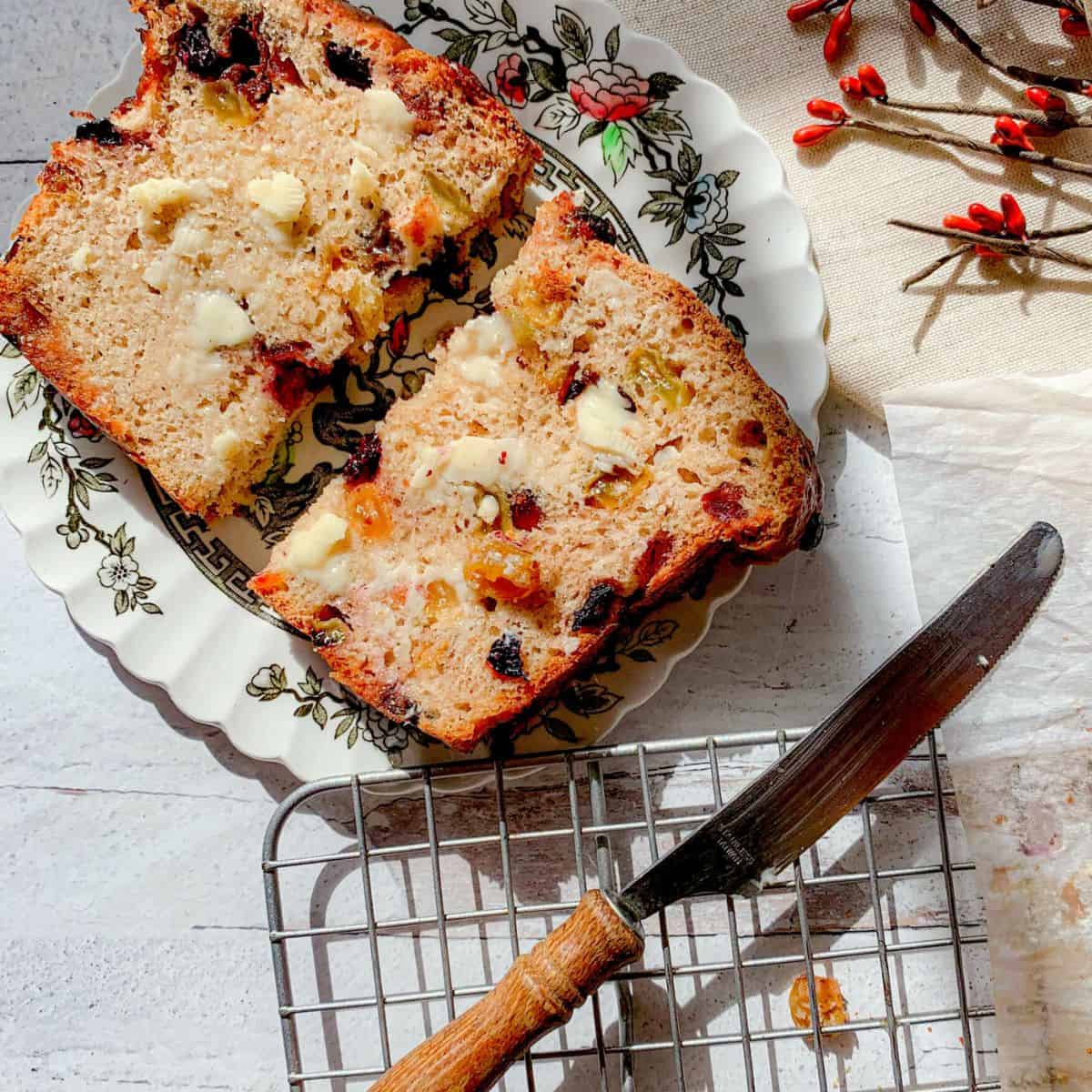 Two slices of banana nut bread with sour cream and butter are on a decorative plate next to a wooden-handled knife and a metal cooling rack; a branch and cloth are in the background.