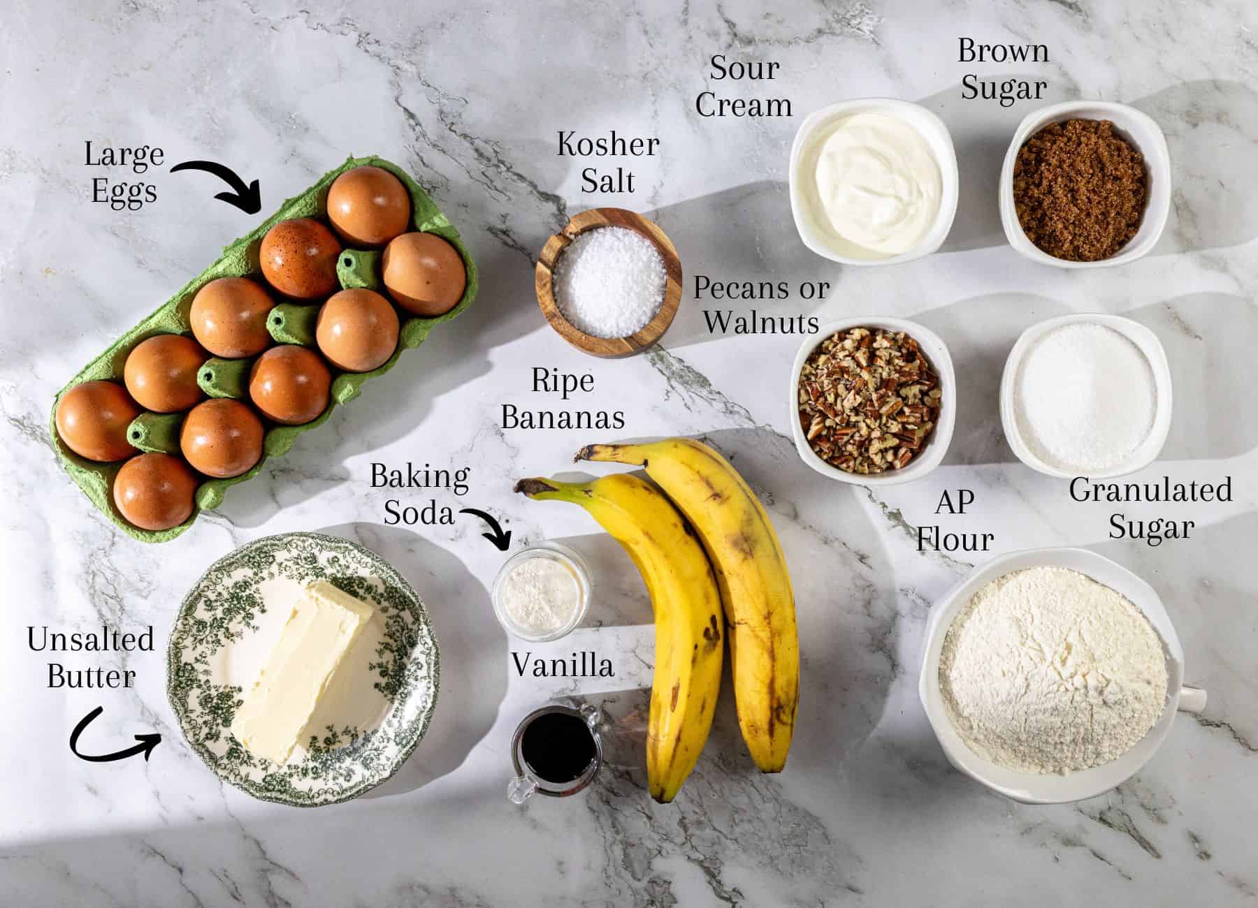 Top-down view of eggs, unsalted butter, baking soda, vanilla extract, ripe bananas, kosher salt, sour cream, chopped pecans or walnuts, brown sugar, granulated sugar, and flour arranged together.