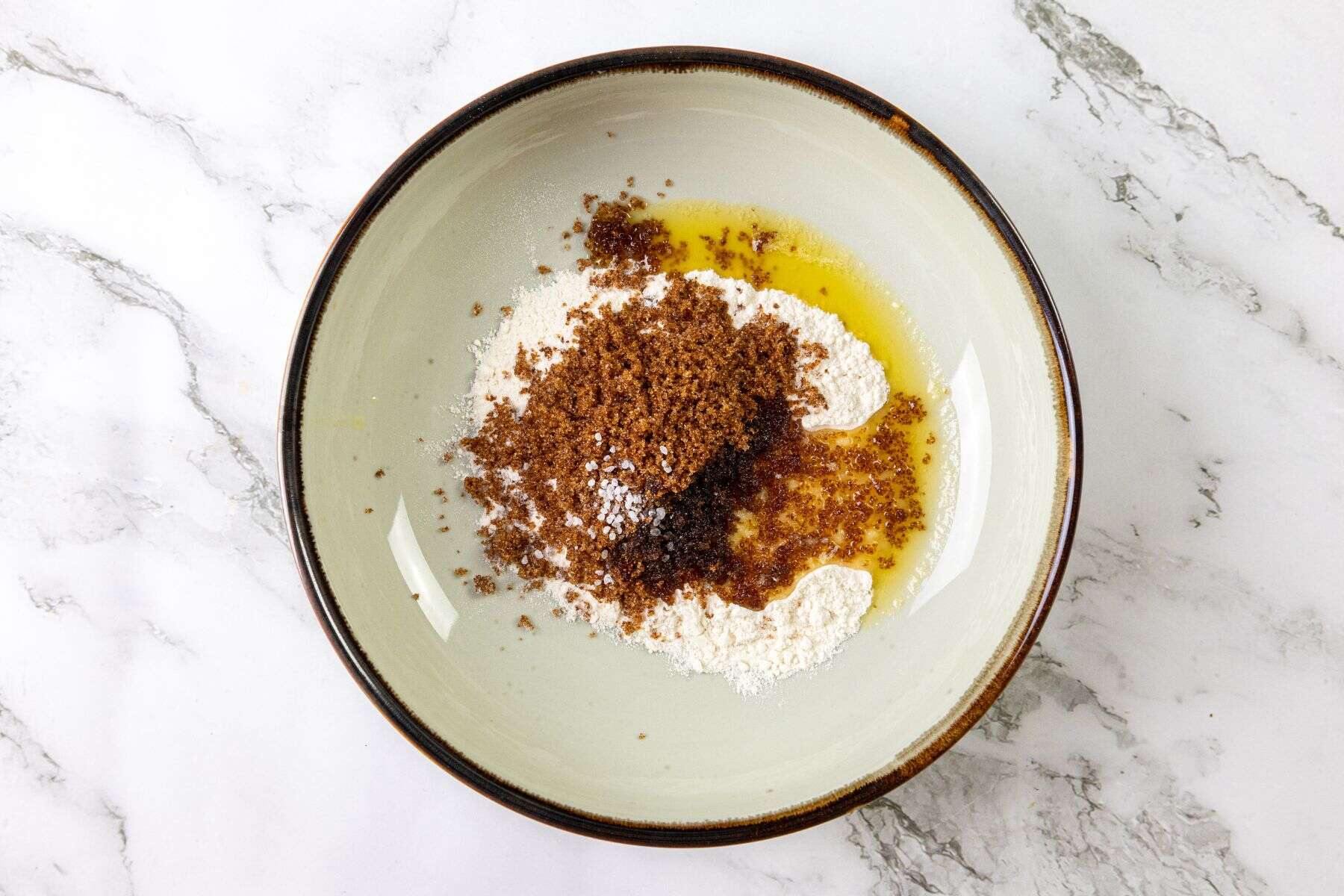A bowl with flour, brown sugar, melted butter, vanilla extract, and sour cream sits on a white marble surface.