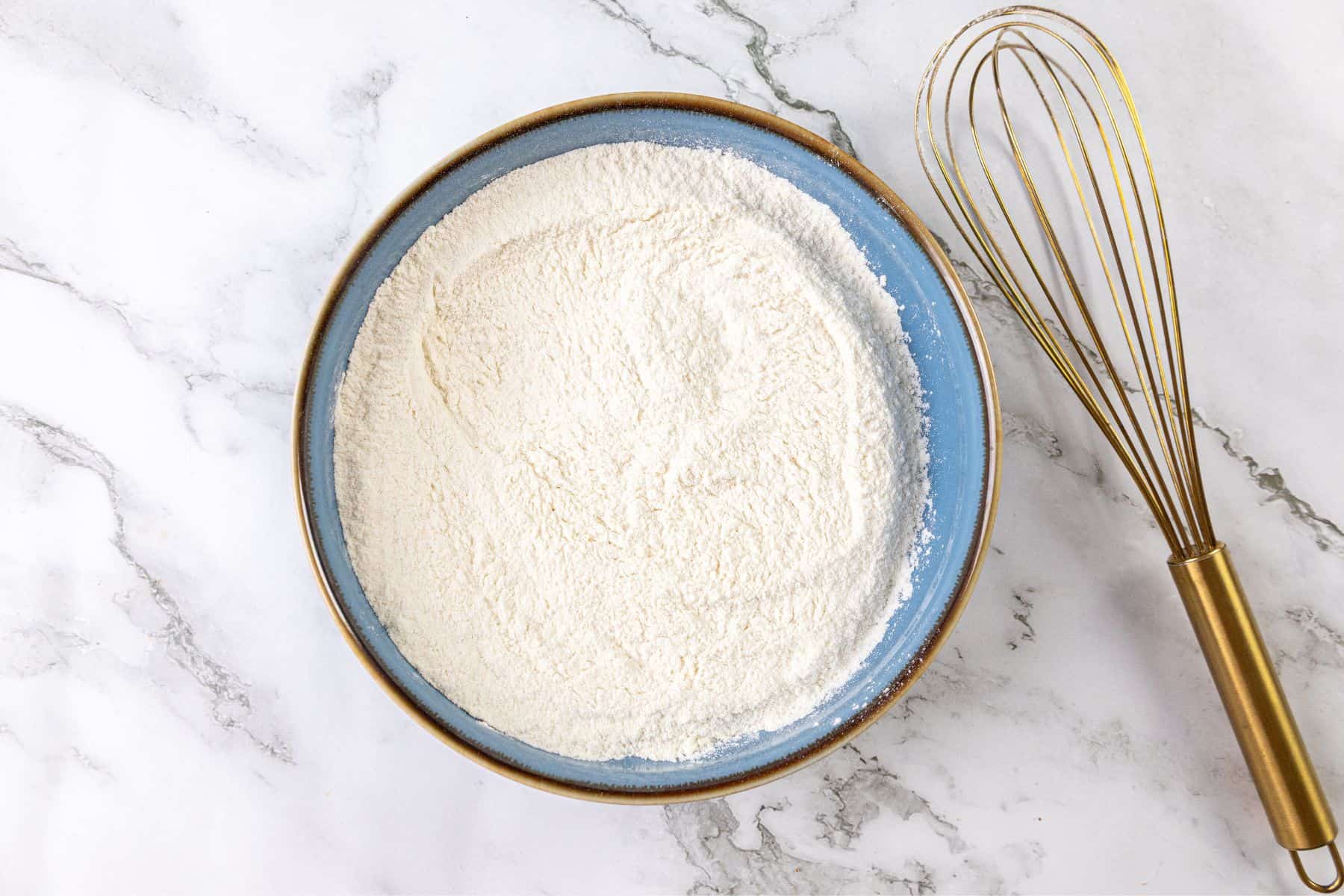 A blue bowl filled with flour sits on a marble surface next to a metal whisk with a gold handle.