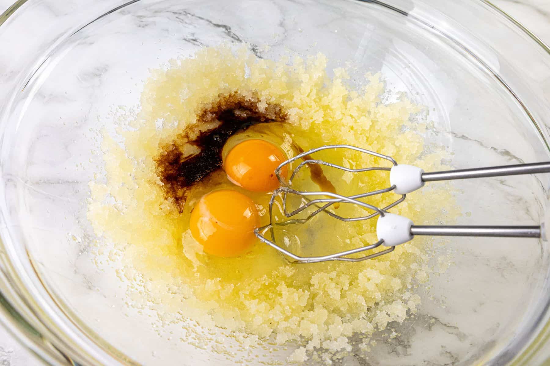 A glass bowl containing sugar, two raw eggs, and vanilla extract being mixed with a hand mixer on a marble surface.