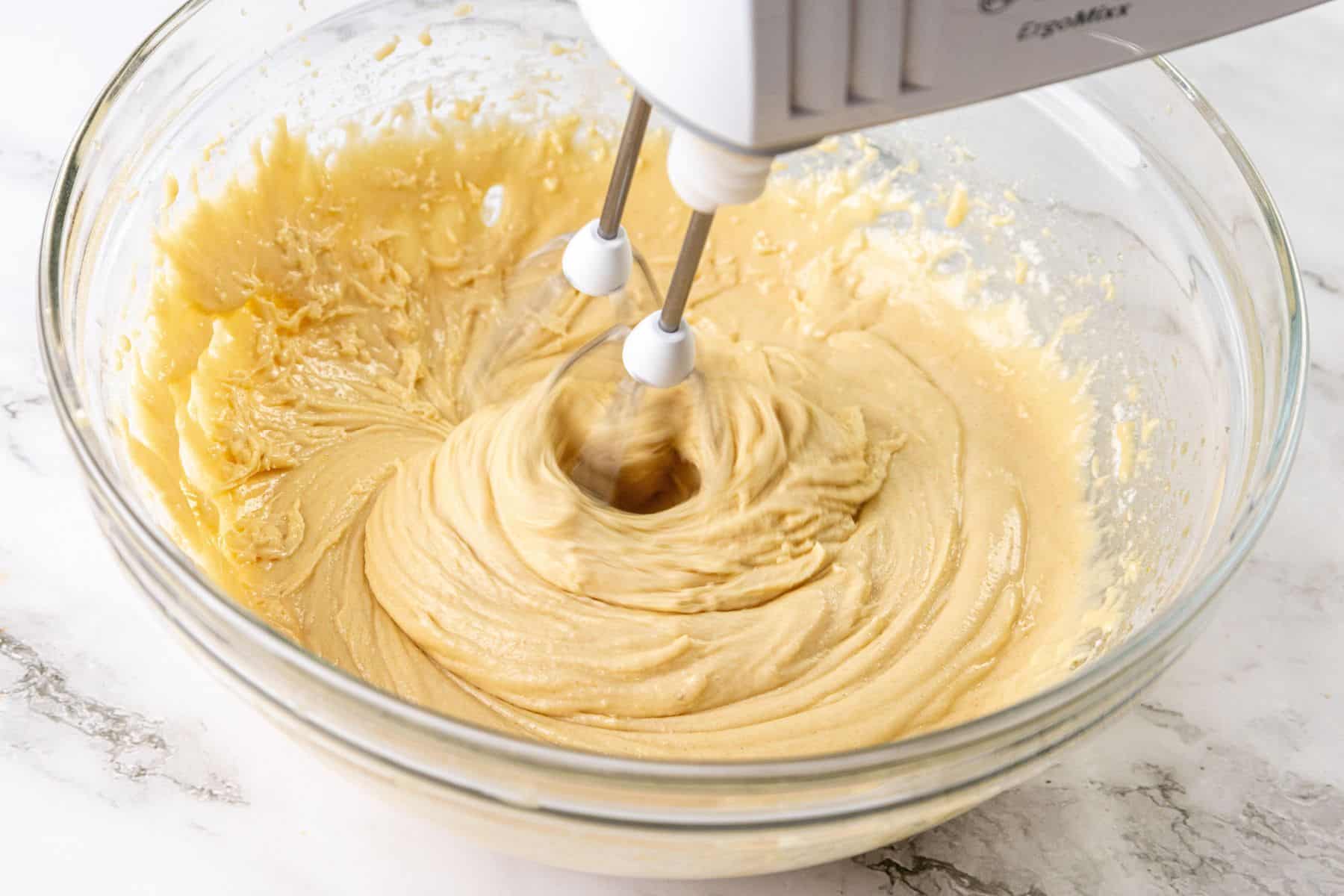 A clear glass bowl containing thick batter with chopped nuts is placed on a marble countertop. An electric hand mixer is partially immersed in the mixture.
