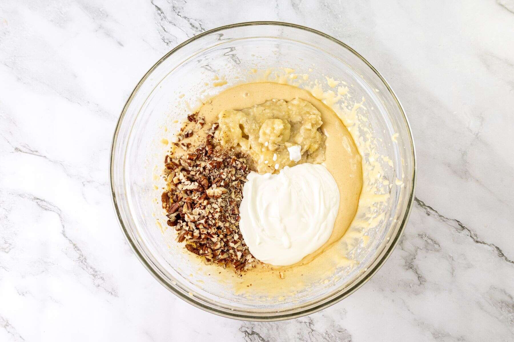 A glass mixing bowl on a marble countertop containing yellow batter, chopped nuts, mashed banana, and sour cream.