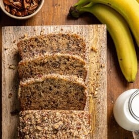 Sliced banana nut bread with chopped nuts on top sits on a wooden board. Whole bananas, a bowl of pecans, and a glass of milk are placed nearby.