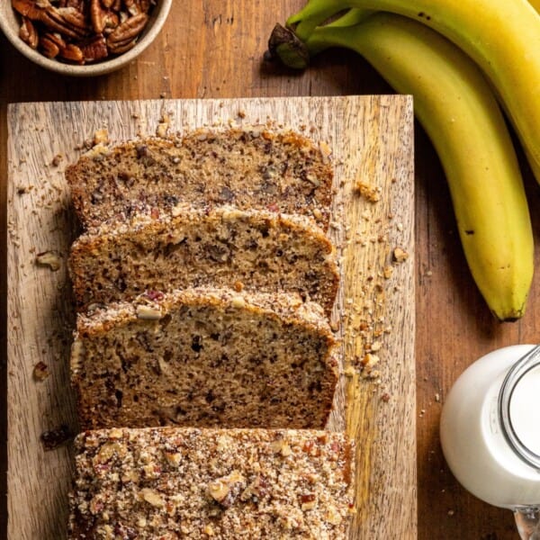 Sliced banana nut bread with chopped nuts on top sits on a wooden board. Whole bananas, a bowl of pecans, and a glass of milk are placed nearby.