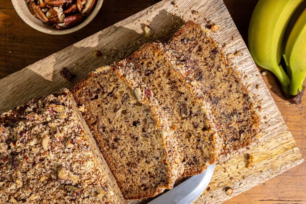 Sliced banana nut bread on a wooden cutting board, accompanied by a bowl of pecans, ripe bananas, and a small dish of sour cream.