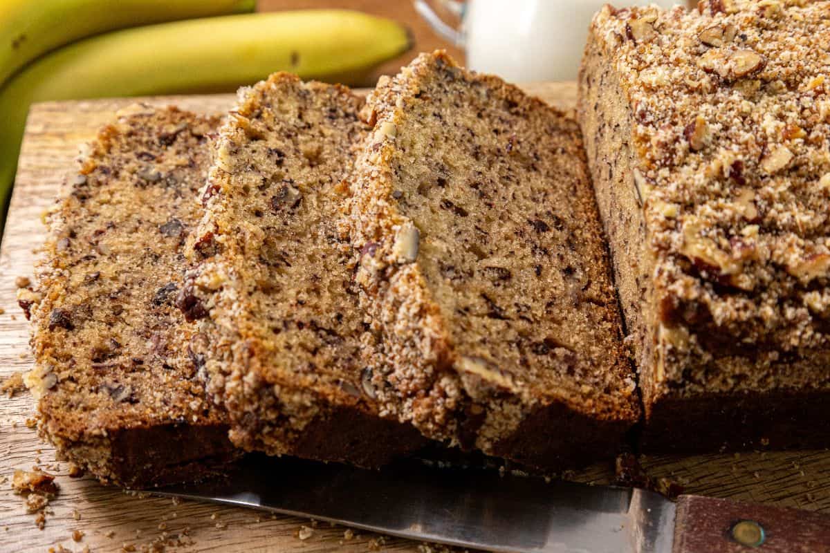 Sliced banana nut bread with a crumb topping on a wooden cutting board, accompanied by a knife, whole bananas, and a glass of milk in the background.