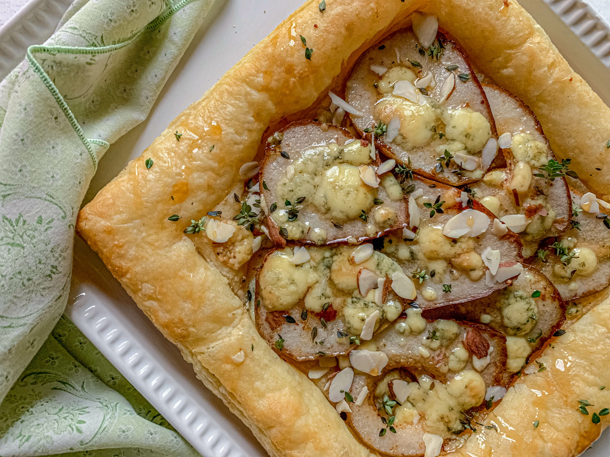 A puff pastry tart topped with sliced pears, crumbled blue cheese, and almond slivers, garnished with fresh thyme. The tart rests on a white plate, accompanied by a green patterned cloth.