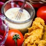 A jar of honey mustard sauce in front of a picnic basket filled with chicken tenders and salad fixings.