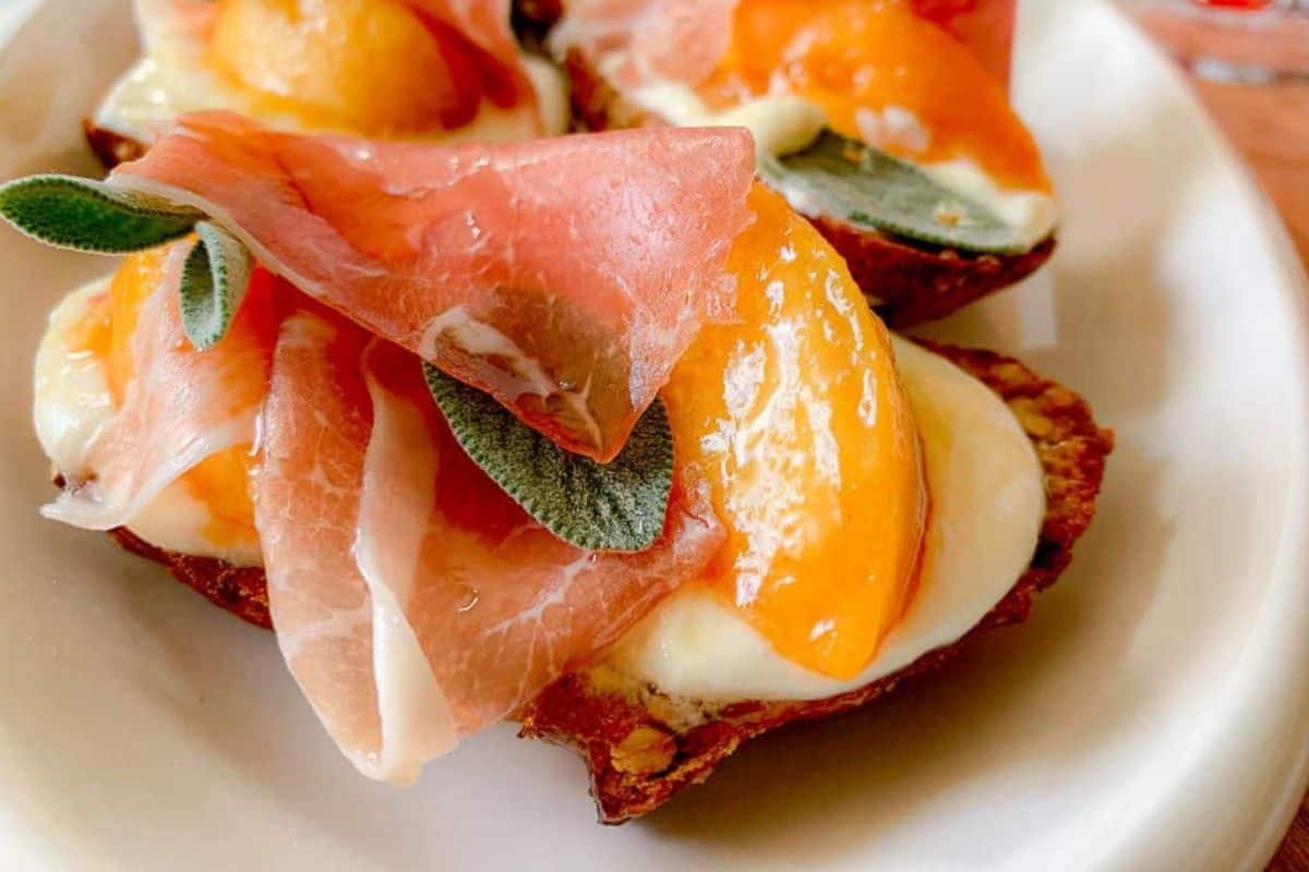 Honey Whipped Ricotta Crostini with Peaches and Prosciutto has honey drizzled over top and is on a serving dish.