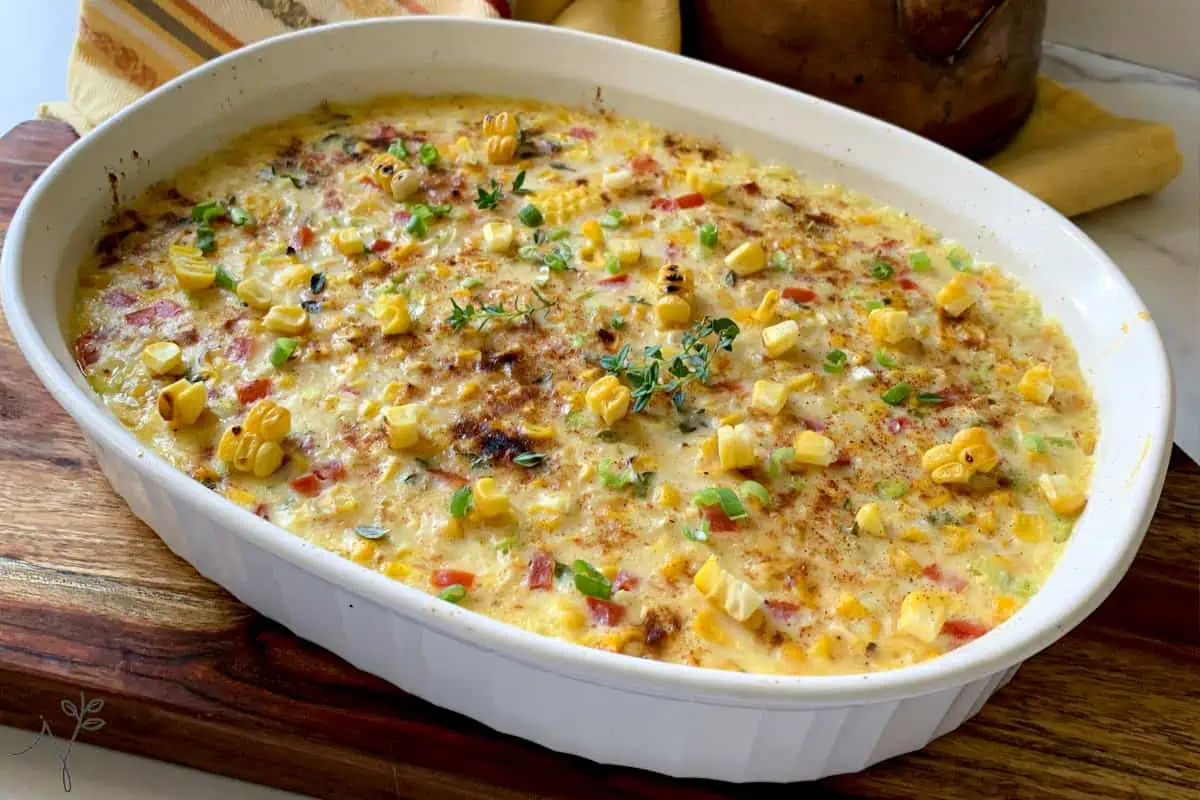 A white rectangular baking dish is filled with a creamy casserole topped with browned cheese, diced red and green bell peppers, corn kernels, and herbs. The dish is placed on a wooden surface, with a yellow napkin in the background.