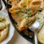 A cast iron pan half filled with a three cheese stuffed shells recipe with creamy cheese sauce and a spoon spooning shells into a bowl.