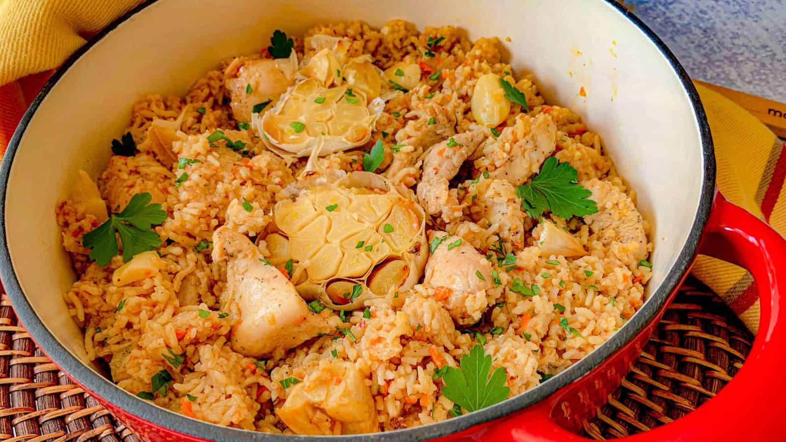 A red pot filled with chicken pieces, rice, whole garlic bulbs, and garnished with chopped parsley.