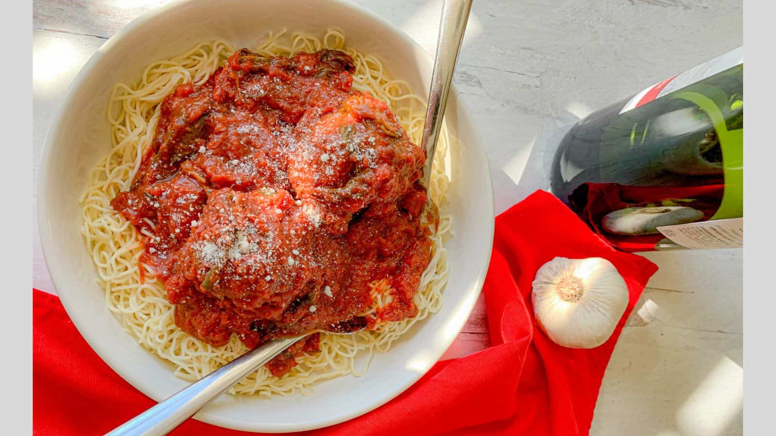 A bowl of spaghetti with red sauce and meatballs on top alongside a head of garlic and a wine bottle.