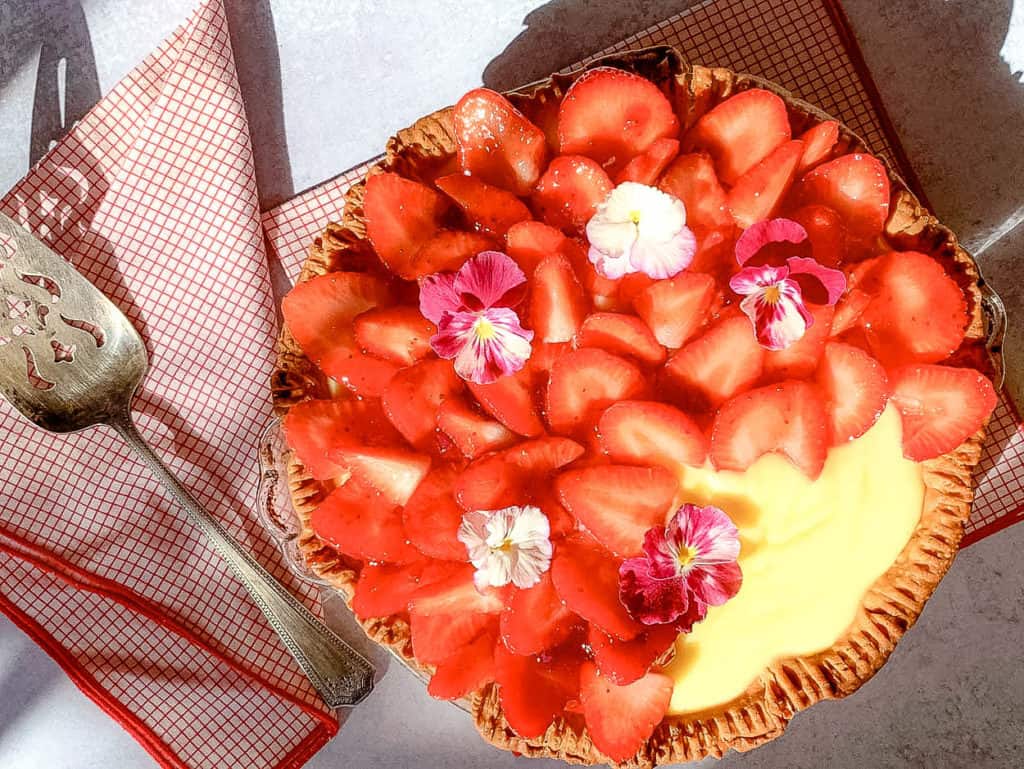 A strawberry tart topped with sliced strawberries and small flowers on a red checkered cloth. A decorative pie server is placed beside it.
