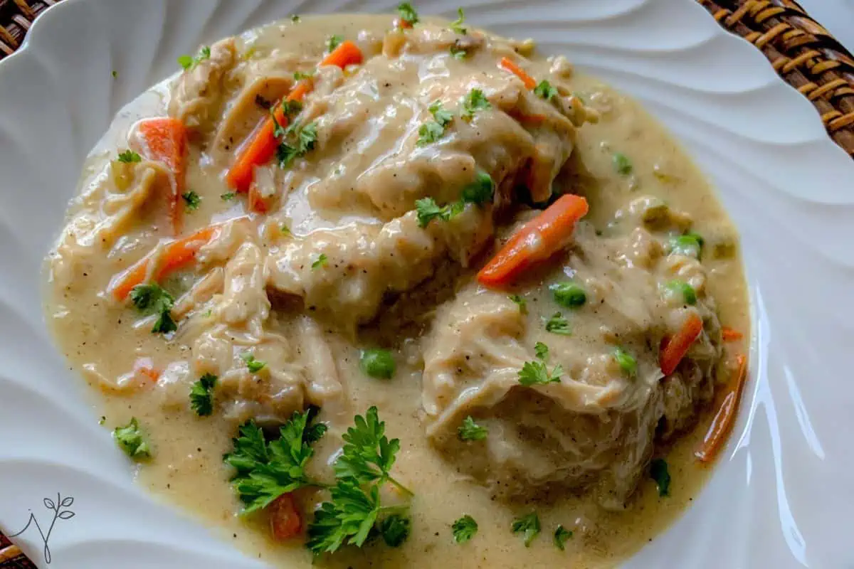 A white plate filled with a creamy stew, featuring chunks of chicken, sliced carrots, peas, and topped with fresh parsley. The dish has a thick, savory sauce and is garnished with green herbs, giving it a hearty and comforting appearance.