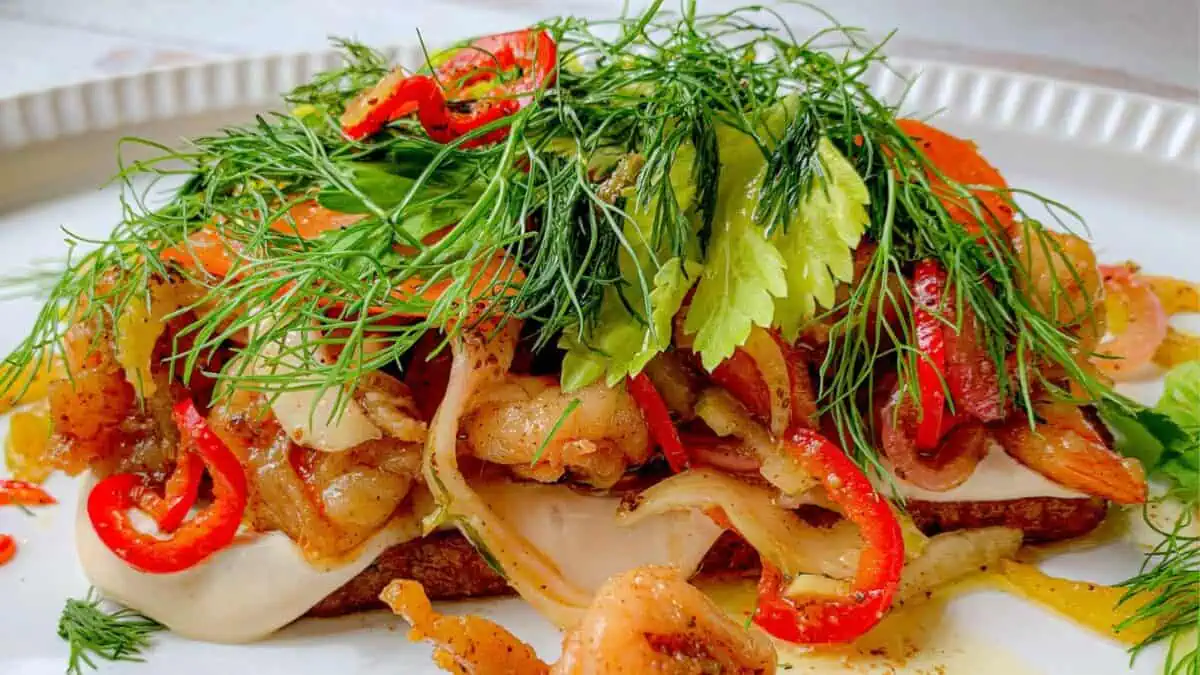 A vibrant open-faced sandwich topped with grilled shrimp, red peppers, caramelized onions, fresh dill, and celery leaves on a white plate. A creamy spread is visible beneath the toppings on a slice of toasted bread.