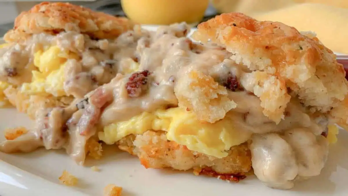 Close-up of a breakfast biscuit sandwich filled with scrambled eggs, crumbled bacon, and topped with creamy sausage gravy. The biscuit is golden brown and flaky, with gravy dripping over the sides onto the white plate. An out-of-focus lemon is in the background.