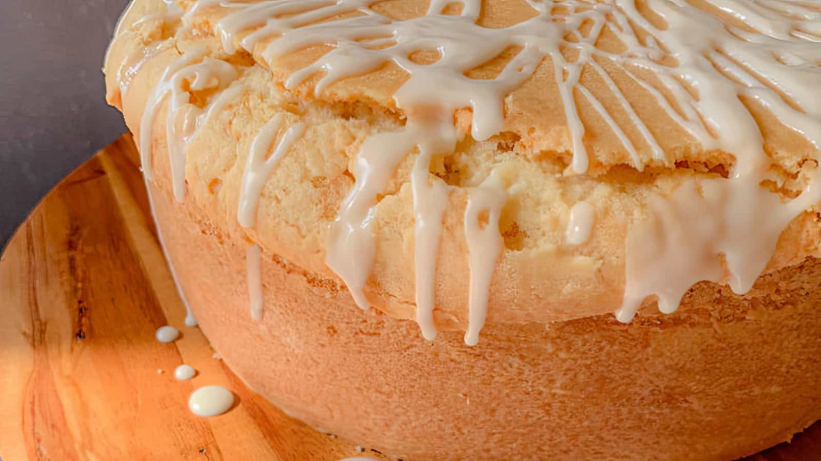 A round, golden pound cake with a cracked top is drizzled with white icing and sits on a wooden surface.