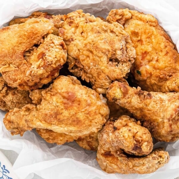 A basket filled with crispy, golden-brown fried chicken pieces, placed on white parchment paper. The background features wooden utensils and a glimpse of mashed potatoes in a dish.