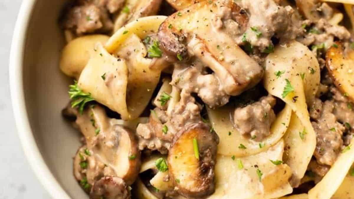 A close-up of wide pasta noodles mixed with ground beef, sliced mushrooms, and a creamy sauce, garnished with chopped parsley.