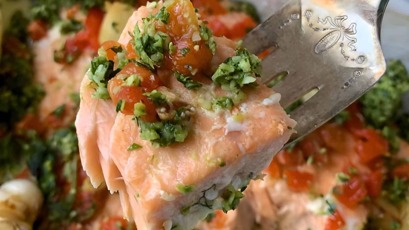 A close-up of a fork holding a piece of cooked salmon topped with chopped herbs and diced tomatoes. The background shows more pieces of salmon garnished similarly with herbs and tomatoes.