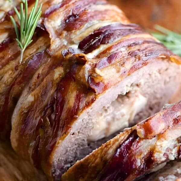 A close-up of a sliced meatloaf wrapped in bacon, garnished with a sprig of rosemary on a wooden cutting board.