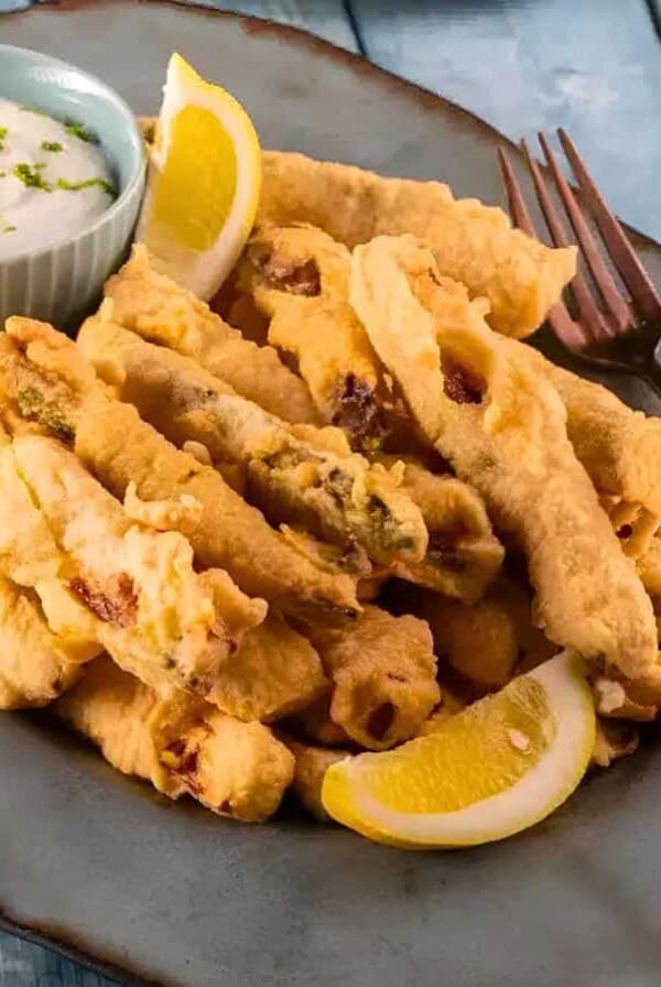 A plate of fried zucchini and squash sticks with lemon wedges, a small bowl of creamy dipping sauce, and a fork on a wooden table.