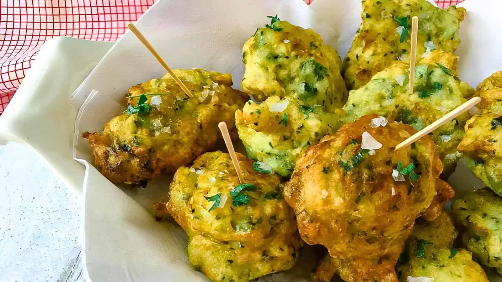 Golden-brown fritters on a plate, garnished with chopped herbs and sea salt, each with a toothpick inserted.