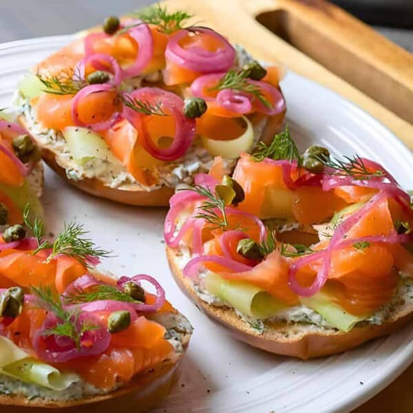 A plate with four bagel halves topped with cream cheese, smoked salmon, pickled red onions, cucumber ribbons, capers, and fresh dill.
