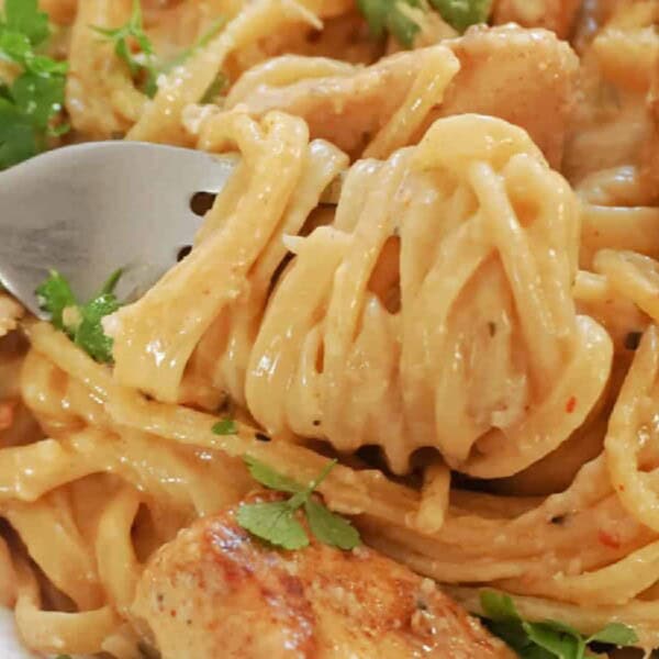 A fork twirls creamy pasta with pieces of seasoned chicken and fresh herbs on a plate.