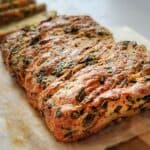 A close-up of a golden, herb-filled cheese bread loaf with melted cheese, resting on parchment paper.