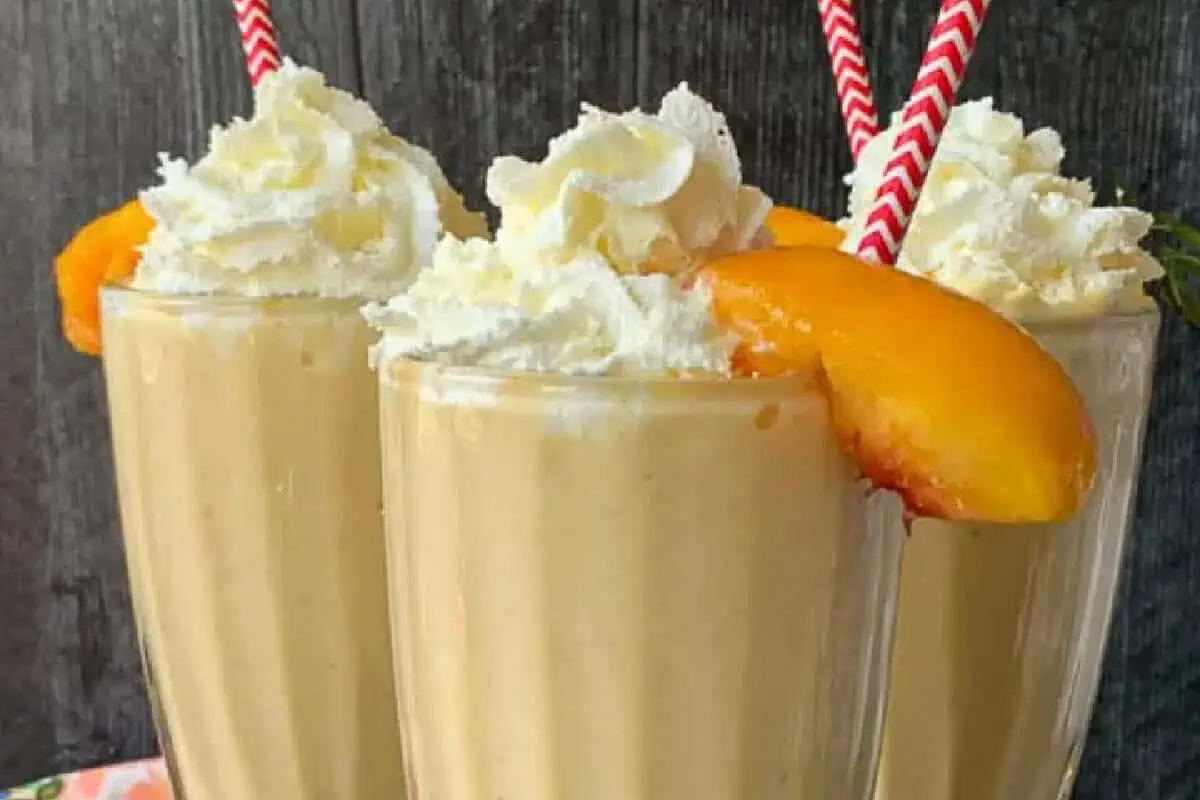 Three tall glasses filled with peach milkshakes, each topped with whipped cream and garnished with a peach slice. Red and white striped straws are inserted in each milkshake. The glasses are arranged against a dark wooden background.