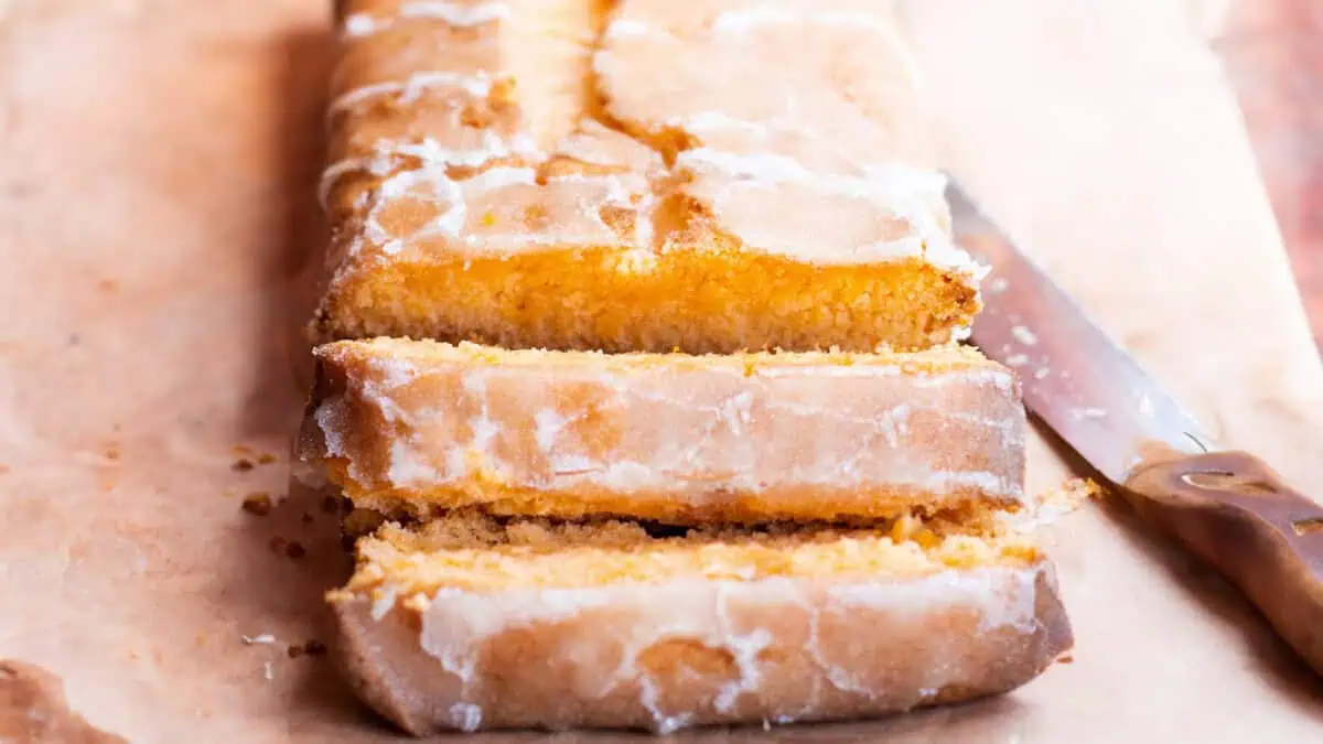 A partially sliced, glazed lemon loaf cake rests on a piece of parchment paper. The moist, yellow cake inside contrasts with the light icing on the outside. A knife with a brown handle lies on the right side of the cake.