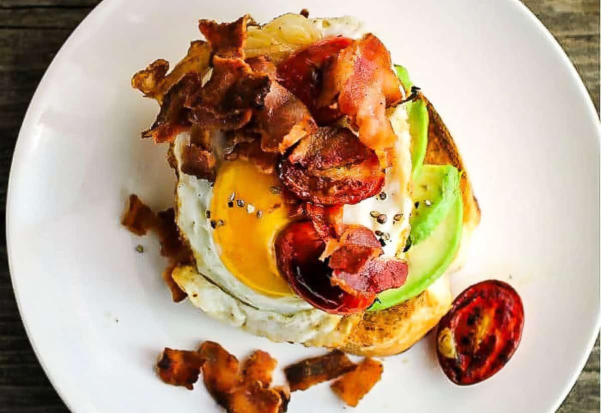 An avocado breakfast toast with bacon, tomatoes, and an egg.