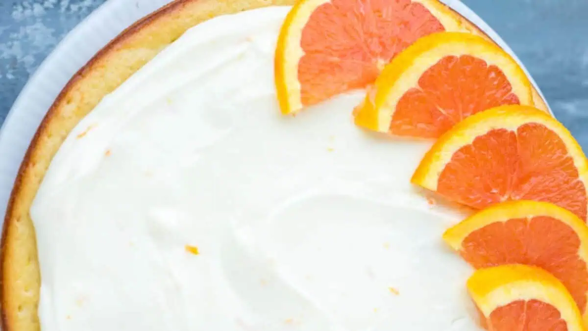 A close-up of a round, frosted cake with five orange slices arranged decoratively in a fan shape on top of the white icing. The cake's edge is slightly visible, and the background features a cool-toned surface.