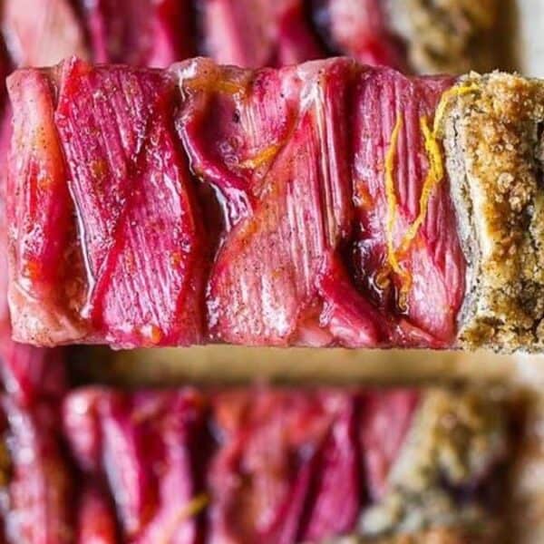 A close-up of a rectangular tart slice being lifted with a spatula, featuring a crumbly crust and glossy, glazed rhubarb pieces on top.