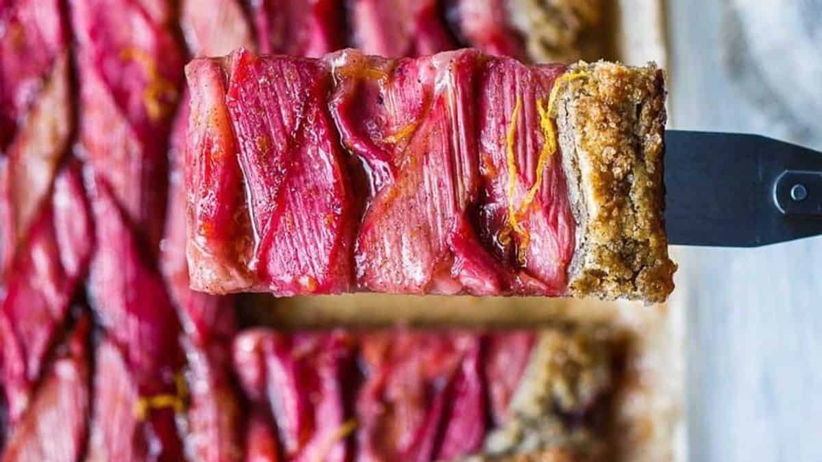 A close-up of a rectangular tart slice being lifted with a spatula, featuring a crumbly crust and glossy, glazed rhubarb pieces on top.