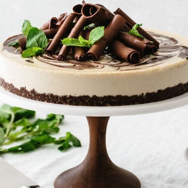 A cheesecake on a wooden cake stand is topped with chocolate curls and mint leaves. In the background, there is a knife, a plate, and fresh mint.