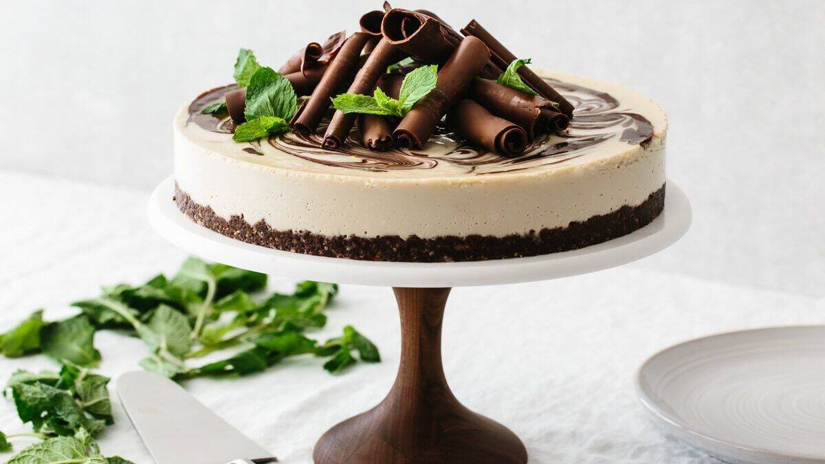 A round cake with a chocolate crust is topped with chocolate curls and mint leaves. It sits on a wooden cake stand, with a plate and knife positioned nearby.