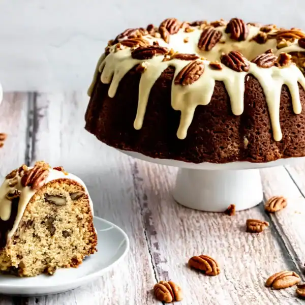 A bundt cake with white icing and pecans on a cake stand, a slice on a plate, and pecans scattered on the wooden surface.