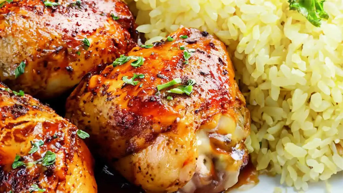 Close-up of glazed chicken thighs garnished with herbs, next to a serving of yellow rice topped with small green herbs.