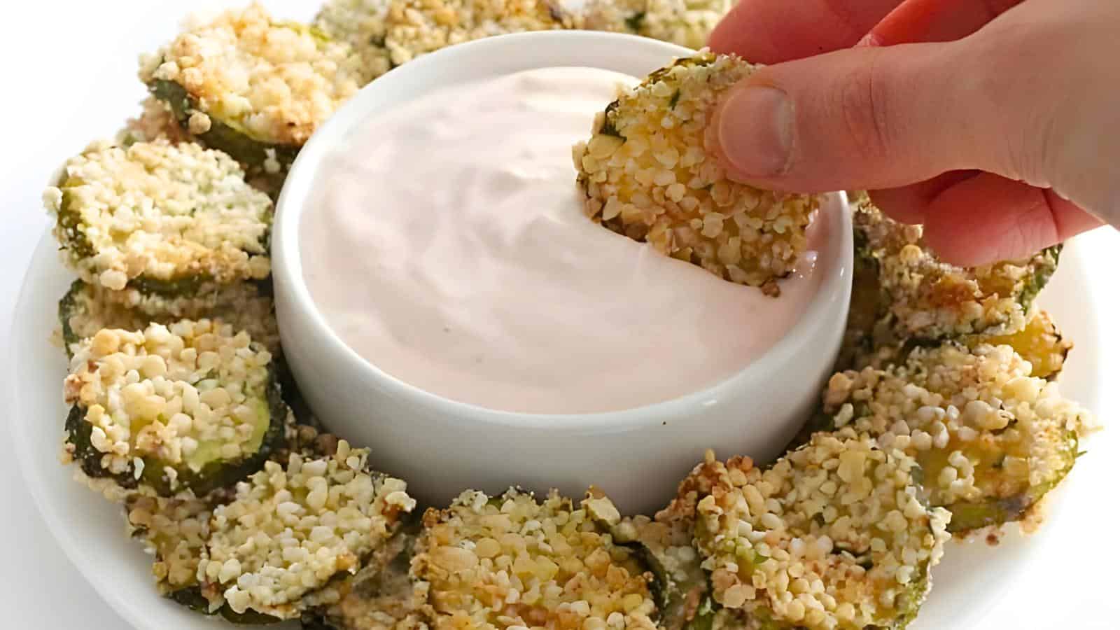 A piece of breaded and fried pickles being dipped into a bowl of creamy dipping sauce, surrounded by more zucchini pieces on a white plate.