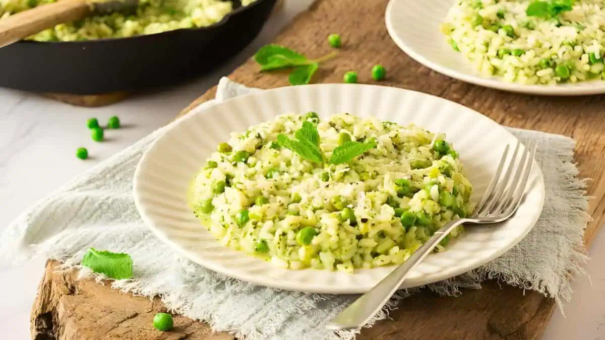 A plate of pea and mint risotto is set on a rustic wooden surface. The creamy rice dish is garnished with fresh mint leaves. A fork is placed on the plate, and there is a skillet with more risotto in the background. Scattered peas add a pop of green to the scene.