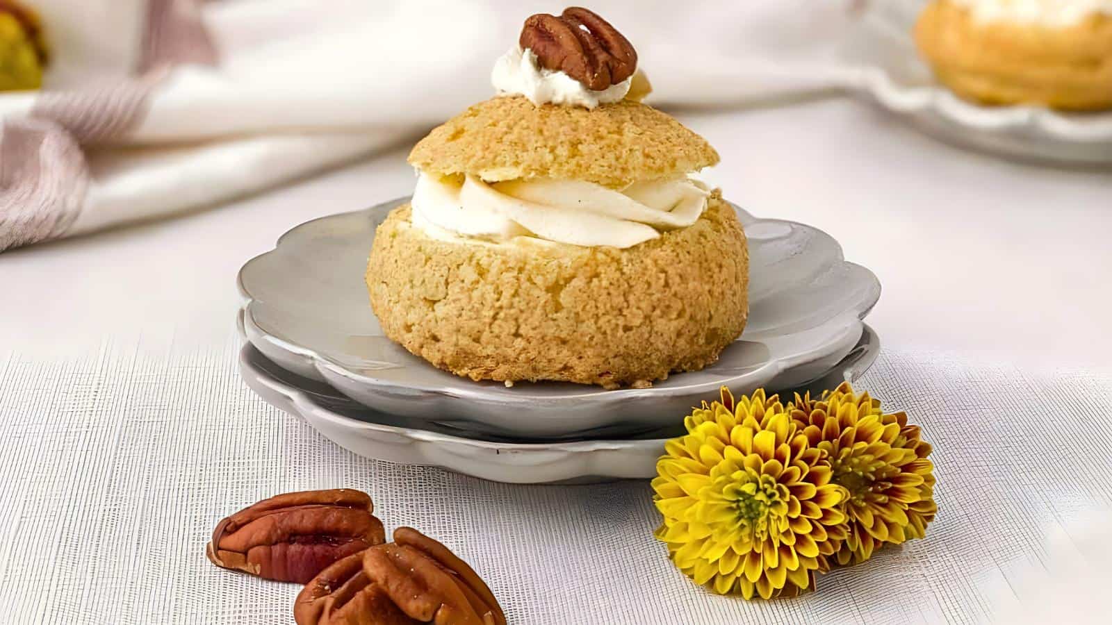 A cream-filled choux pastry topped with a pecan sits on a gray ceramic plate, accompanied by a yellow chrysanthemum flower and pecan halves.
