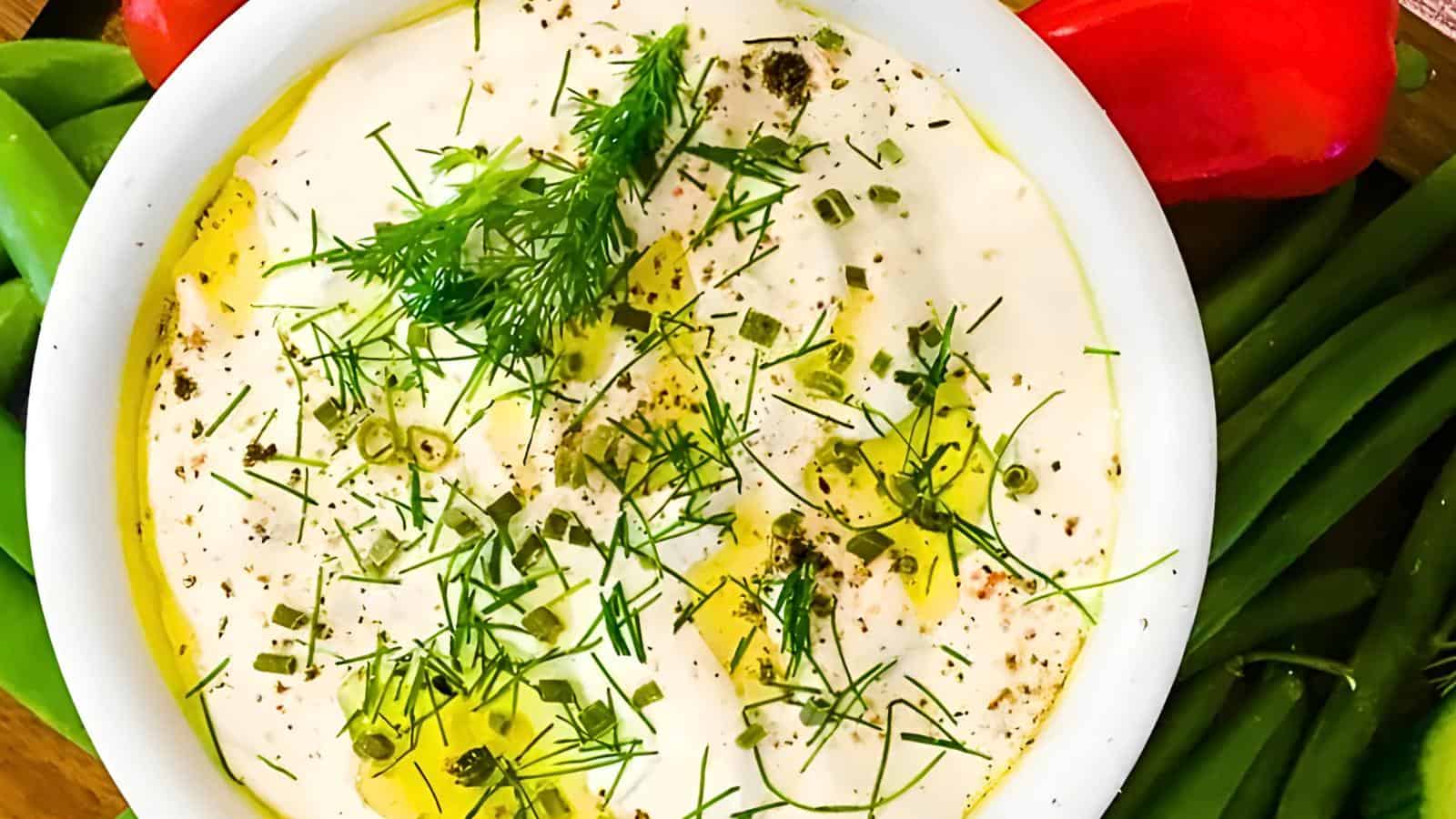 A bowl of white dip garnished with chopped herbs, surrounded by green beans and red vegetables.