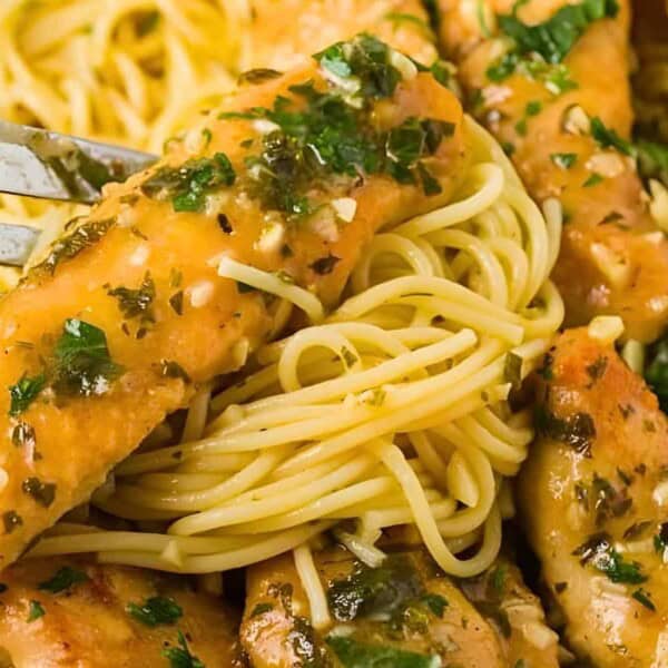 A fork holding a piece of cooked chicken and spaghetti, garnished with herbs and a savory sauce. Chicken breasts over a bed of spaghetti, visible garlic, and parsley.